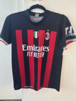 AC Milan matchtröja R. Leão 17 - Snygg AC Milan matchtröja med R. Leão och nummer 17 på ryggen. Tröjan är svart med röda ränder, klubbmärke och sponsortryck framtill. Ärmarna har italienska flaggans färger i mudden. Tillverkad i lätt och ventilerande polyester, perfekt för fotbollsträning.