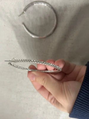 Stora silvriga hoops med pärldetaljer - Säljer ett par stora silvriga hoop-örhängen med glittrande pärldetaljer längs hela ringen. Örhängena är tunna och har en klassisk rund form som ger en snygg och trendig look. Perfekta för att lyfta vilken outfit som helst.