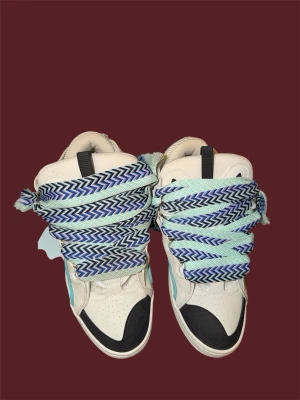 Vita high top sneakers med mönstrade snören - Unika vita high top sneakers med svarta detaljer och turkosa inslag. Skorna har extra breda, mönstrade skosnören i blått och ljusgrönt zigzag-mönster. Ovandelen är i läder och textil, med vadderad krage och svart hälflik. Perfekta för dig som vill sticka ut.