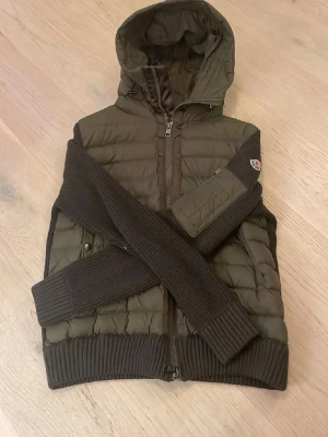 Olivgrön Moncler Cardigan  - Snygg olivgrön cardiganjacka från Moncler med quiltad framsida och huva. Ärmar och rygg är stickade i matchande mörkgrönt material. Jackan har dragkedja framtill, ribbade muddar och två fickor med knapp. Perfekt för dig som gillar streetwear och exklusiva detaljer.