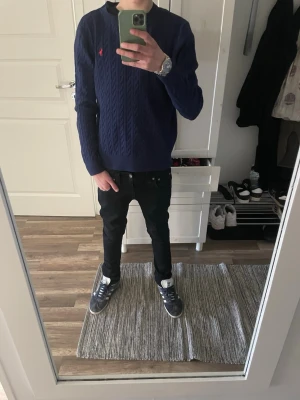 Mörkblå kabelstickad tröja Polo Ralph Lauren - Snygg mörkblå kabelstickad tröja från Polo Ralph Lauren med rund halsringning och röd broderad logga på bröstet. Tröjan har långa ärmar och ribbade muddar vid ärmslut och nederkant. Perfekt till jeans för en clean och klassisk look. Kan gå ner i pris vid en snabb affär!
