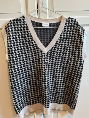 Svartvit rutig väst från VILA - Snygg stickad väst från VILA i svartvitt rutigt mönster. Västen har v-ringning och ribbade kanter i beige. Perfekt att styla över en skjorta eller t-shirt för en trendig look.