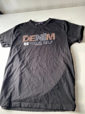 Svart t-shirt med DENIM-tryck - Svart t-shirt från Denim med trycket 'DENIM BE YOUR SELF' i vitt och brunt på bröstet. Klassisk rund hals och korta ärmar. Tillverkad i mjuk bomull, perfekt för en avslappnad stil.
