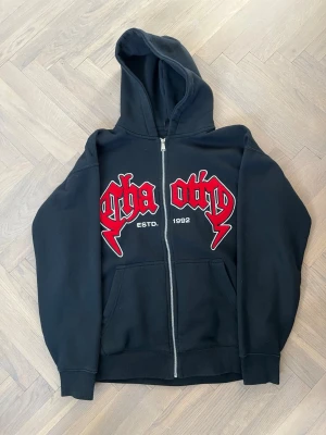 Svart zip hoodie med röd text - Säljer en svart hoodie med dragkedja från Otha. På bröstet finns stor röd text i gotisk stil och detaljer som 'ESTD. 1992'. Hoodien har huva och två fickor framtill. Perfekt för dig som gillar streetwear och vill sticka ut.