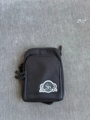 Svart axelväska med tryck - Säljer en svart axelväska i tyg med ett coolt tryck framtill. Väskan har dragkedja upptill och en extra ficka på framsidan. Perfekt för att bära det viktigaste och enkel att matcha med olika outfits.