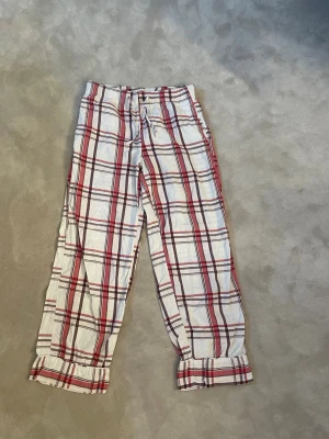 Pyjamasbyxor Gina  - Säljer ett par klassiska pyjamasbyxor i vitt med rött och brunt rutmönster. Byxorna har resår och dragsko i midjan samt rynkade benslut för extra komfort. Perfekta för mysiga hemmakvällar.