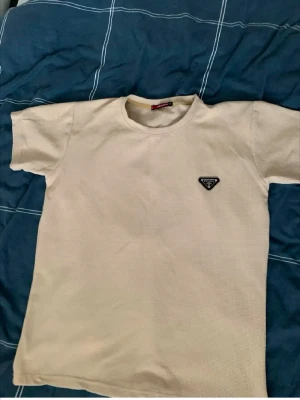 Beige t-shirt från Prada - Säljer en beige t-shirt i storlek XL. T-shirten har korta ärmar, rund halsringning och ett diskret märke på bröstet. Materialet är bomull med en strukturerad, ribbad känsla som ger en snygg och modern look.