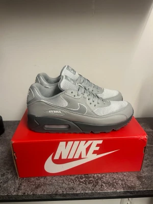 Nike Air Max 90 grå sneakers - Snygga Nike Air Max 90 sneakers i helgrå färg med klassisk siluett. Ovandelen är i mesh och syntet med ikonisk Air Max-dämpning i sulan. Tidlös design med vita detaljer och robust yttersula. Perfekta för dig som gillar streetwear och sportig stil.