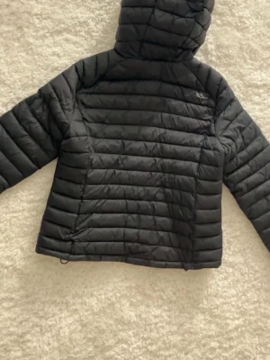 Svart dunjacka med huva från Peak Performance - Snygg svart dunjacka från Peak Performance med huva och dragkedja framtill. Jackan har quiltad design och två fickor med dragkedja. Perfekt för kalla dagar och enkel att matcha med olika outfits.