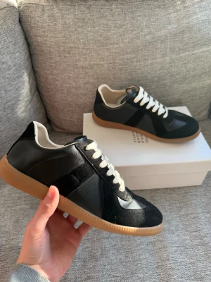 Svarta sneakers från Maison Margiela - Snygga svarta sneakers från Maison Margiela med vita skosnören och beige gummisula. Skorna har en mix av mocka och läderdetaljer, samt en klassisk låg siluett. Perfekta för dig som gillar stilrena och tidlösa sneakers. Bara att höra av er om ni har några frågor, funderingar eller vill ha flera bilder 🙌