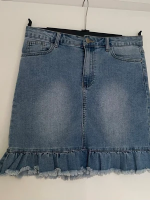 Blå jeanskjol med volangkant - Säljer en blå jeanskjol med volang och fransad kant längst ner. Kjolen har klassiska fickor fram och bak, knapp och dragkedja i midjan. Perfekt för dig som gillar denim och vill ha en cool, trendig look.