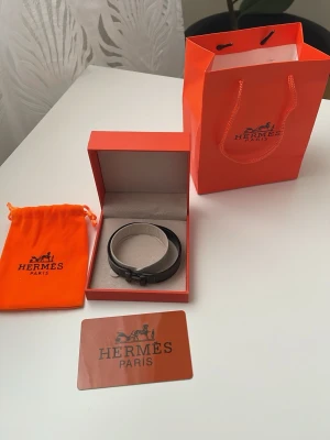 Svart armband i läder - Snyggt svart armband i slätt läder med metallspänne format som ett H. Kommer i originalförpackning med orange påse och box. Perfekt accessoar för dig som gillar stilrena och exklusiva detaljer.