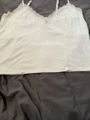 Vit linnetopp med spets - Söt vit linnetopp med tunna axelband. Toppen har spetskant längs urringningen och är tillverkad i mjukt bomullsmaterial. Perfekt för att addera lite extra till din outfit.