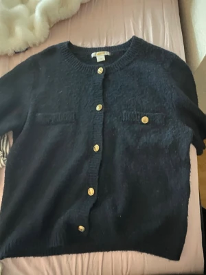 Marinblå kofta från Gina Tricot - Stilren marinblå kofta från Gina Tricot med guldfärgade knappar och två dekorativa bröstfickor. Koftan har rund halsringning och ribbade muddar. Perfekt att slänga över en topp för en snygg lager-på-lager-look.