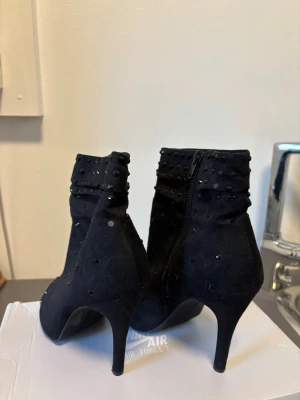 Svarta stövletter med nitar och öppen tå - Snygga vintage svarta stövletter med 10 cm och öppen tå. Skorna är dekorerade med små svarta ”kristaller” över hela ytan (någon har fallit av) och har dragkedja på sidan. Tillverkade i mockaimitation för en mjuk känsla och trendig look.