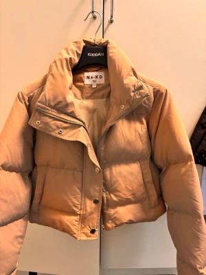 Beige kort pufferjacka från NA-KD - Säljer en beige, kort pufferjacka från NA-KD med hög krage och dragkedja samt tryckknappar framtill. Jackan har en boxig passform och breda kanaler som ger en riktigt puffig look. Perfekt för kalla vinterdagar och enkel att matcha med olika outfits.