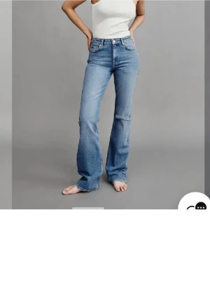 Blå bootcut jeans med slitning - Säljer ett par blå bootcut jeans med hög midja och lätt slitning på ena benet. Jeansen har klassisk femficksdesign och är tillverkade i denim. Perfekt för dig som gillar en avslappnad och trendig look.