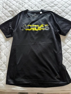Svart Adidas t-shirt med tryck - Snygg svart t-shirt från Adidas med korta ärmar och ett stort grafiskt Adidas-tryck i svart grått och gult över bröstet. Tillverkad i ett lätt och sportigt polyestermaterial som passar perfekt till träning eller chill. Adidas-logga även nere vid fållen. Bra skick