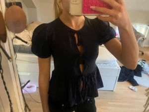 Svart blus med puffärm och knyt - Säljer en svart blus med puffärmar och flera knyt framtill. Blusen har en lätt utsvängd nederdel och är tillverkad i ett mjukt tyg. Perfekt för dig som gillar unika detaljer och en feminin siluett. Storlek 146/152