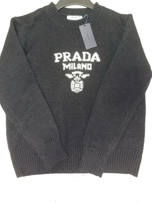 Prada Milano tröja - Snygg svart  stickad tröja från Prada med vit logotyp och texten 'PRADA MILANO' på bröstet. Tröjan har rund halsringning, ribbade muddar vid ärmslut och nederkant samt en klassisk passform. Perfekt för dig som gillar exklusiva märken och stilren design. Helt ny! Går bra att buda ! Jag beställde stl S och tröjan är storlek S