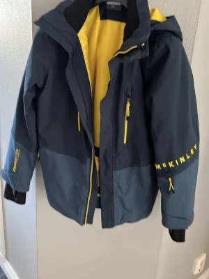 Mörkblå fodrad jacka  McKinley stl 158 - Snygg mörkblå vinterjacka från McKinley med gula detaljer och dragkedjor. Jackan har huva, justerbara ärmslut och en gul insida. Fodrad med snödroppar på insidan och vid ärmslut. Stl 158