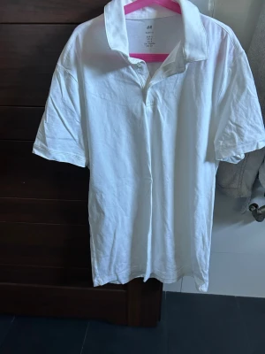 Vit pikétröja från H&M - Klassisk vit pikétröja från H&M med krage och korta ärmar. Tillverkad i mjuk bomull som känns skön mot huden. Enkel och stilren design som passar till många olika looks.