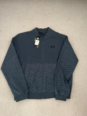 Grå zip-jacka från Under Armour - Sportig grå zip-jacka från Under Armour med diskret logga på bröstet och mönstrade partier med texten 'Under Armour' på ärmar och nedre delen. Tillverkad i ett lätt och funktionellt material, perfekt för träning eller chill. Ribbstickade muddar och hög krage. PRIS GÅR ATT DISKUTERAS 