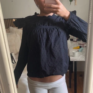 Svart blus med volang från ONLY - Trendig svart blus från ONLY med volangdetaljer vid halsen och axlarna. Blusen har långärmad design och är gjord i ett lätt, strukturerat tyg med diskret prickmönster. Perfekt för dig som vill ha en stilren men ändå unik look.