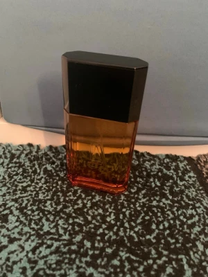 Azzaro Amberfärgad Parfymflaska - Snygg parfymflaska från Azzaro i en stilren, rektangulär design. Flaskan är gjord av glas med en snygg övergång från bärnstensfärgat till rött och har ett svart, kantigt lock. Perfekt för dig som gillar eleganta och moderna doftflaskor.