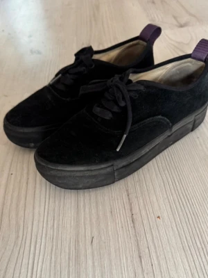 Svarta Eyty’s sneakers med platåsula - Snygga svarta sneakers med tjock platåsula och ovandel i mocka. Skorna har svarta skosnören och en lila hälflik för enkel påtagning. Perfekta för dig som gillar en clean och stilren look med lite extra höjd. Använda men har mycket kvar att ge!