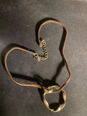 Brunt halsband med vriden guldring - Unikt halsband med ett brunt mockaband och en vriden ring i guldfärgad metall som hänge. Justerbart med kedja och karbinlås. Perfekt för dig som gillar en bohemisk och stilren look.OBS! Materialet är okänt, kan ej garantera att det är rostfritt.