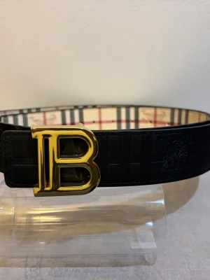 Svart Burberry bälte med guldspänne - Snyggt svart bälte från Burberry med ett stort guldigt B-spänne. Insidan har det klassiska rutiga Burberry-mönstret i beige, rött och svart. Perfekt accessoar för att lyfta vilken outfit som helst. Funkar på båda sidorna vilket gör den till en tvåsidig bälte