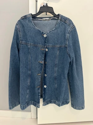Blå jeansjacka med rund hals Gina tricot - Snygg blå jeansjacka med rund halsringning och långa ärmar. Jackan har en rak passform och stängs med silverfärgade knappar framtill. Klassisk sömstruktur och utan krage för en modern look. Perfekt att slänga över en hoodie eller t-shirt.