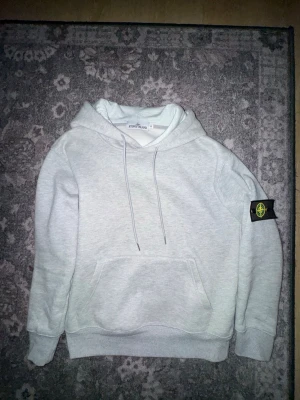 Ljusgrå hoodie från Stone Island - Säljer en stilren ljusgrå hoodie från Stone Island med klassisk huva och dragsnören. Hoodien har en stor magficka och den ikoniska Stone Island-loggan på vänster ärm. Perfekt för dig som gillar streetwear och vill ha något bekvämt och snyggt.