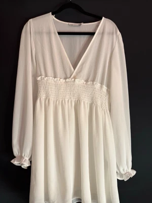 Vit kort omlottklänning med v-ringning - Söt vit kortklänning med v-ringning och omlott framtill. Klänningen har långa ärmar med resår vid ärmslut och smockad midja som ger en fin siluett. Tillverkad i ett lätt, luftigt material som passar perfekt till varmare dagar.