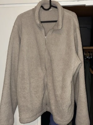 Beige teddyjacka med dragkedja - Mysig beige teddyjacka med hel dragkedja framtill och hög krage. Jackan har en avslappnad passform och är tillverkad i fluffigt teddy-material som håller dig varm. Perfekt för kyliga dagar när du vill ha något både bekvämt och stilrent.