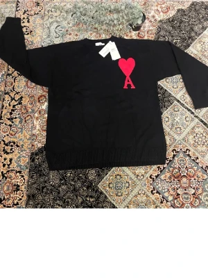 Svart stickad tröja från Ami - Svart stickad tröja från Ami Alexandre Mattiussi med röd hjärt- och A-detalj på bröstet. Tröjan har rund halsringning och ribbstickade muddar vid ärmslut och nederkant. Perfekt för dig som gillar stilrena plagg med en twist.