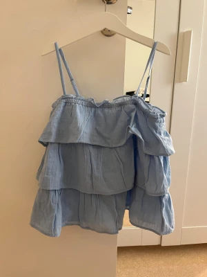 Ljusblå volangtop med smala band - Supersöt ljusblå sommartopp från Gina tricot  med tunna axelband och flera lager volanger. Tillverkad i mjuk bomull som känns luftig och skön. Perfekt för dig som gillar en feminin och lekfull stil.