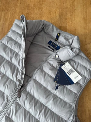 Ljusgrå dunväst från Polo Ralph Lauren - Snygg ljusgrå dunväst från Polo Ralph Lauren med klassisk ståkrage och dragkedja framtill. Västen har två sidofickor med dragkedja och den ikoniska Polo-loggan broderad på bröstet. Perfekt för lager-på-lager och stilren look.