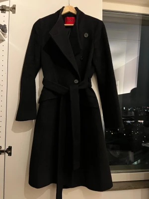 Svart ullkappa från Hugo Boss - Stilren svart kappa från Hugo Boss med markerad midja och knytskärp. Kappan är gjort av ull & jungfruull. Kragen kan knäppas hela vägen eller ”ligga” ner som en vanlig kappa. 