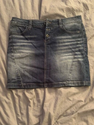 Blå jeanskjol med knappar - Snygg blå jeanskjol i klassisk modell med framfickor, bakfickor och knappgylf. Kjolen har bälteshällor och är i en kort längd, perfekt till vardagslooken. Materialet är slitstarkt denim som ger en cool och avslappnad vibe.