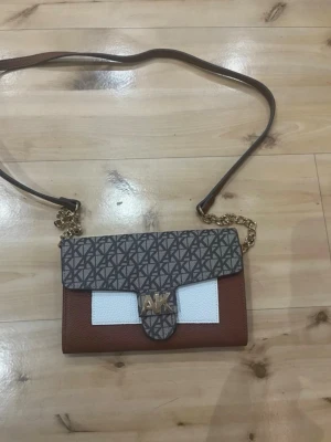 Brun och vit axelremsväska AK - Snygg axelremsväska från AK med brunt och vitt konstläder, guldfärgad kedjedetalj och magnetlås. Väskan har ett grafiskt mönster på locket och flera fack inuti, inklusive dragkedjefack och kortfack. Perfekt för dig som vill ha både stil och funktion.