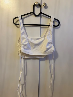 Vit croppad linnetopp med snörning - Trendig vit croppad linnetopp med breda axelband och snörning i sidorna för justerbar passform. Toppen har en ribbad struktur och är perfekt för varma dagar. Enkel att matcha med både jeans och kjol.