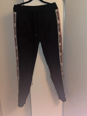Svarta Moschino mjukisbyxor - Säljer ett par svarta mjukisbyxor från Moschino med vita sidor och logga längs båda benen. Byxorna har dragsko i midjan och smal passform med muddar vid bensluten. Perfekta för en chill och trendig look. Byxorna är storlek S, kvitto medföljer. 