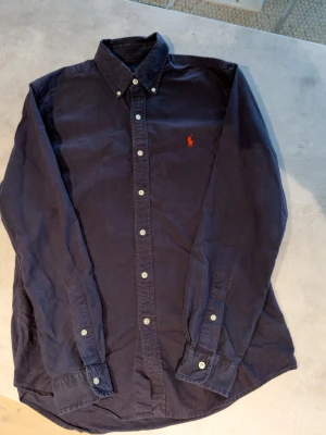 Mörkblå skjorta från Ralph Lauren - Marinblå skjorta från Ralph Lauren med röd loga i vuxenstorlek M och väl omhändertagen dessutom i bra skick, bara att höra av sig vid funderingar. Kan skickas inom en dag!