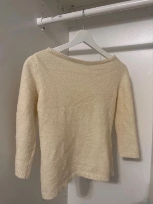 Krämvit stickad tröja med glitter - Mysig krämvit stickad tröja med diskret glittereffekt. Tröjan har trekvartsärmar, rund halsringning och ribbade muddar. Perfekt för dig som gillar en enkel men ändå lite extra look.