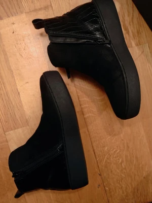 Svarta stövletter med dragkedja storlek 38 Nyskick - Snygga svarta stövletter med dragkedja på sidan och detaljer i både mocka och glansigt material med krokopräglad look. Skorna har en rund tå och platt sula, vilket ger en cool och modern vibe. Perfekta för dig som vill ha en stilren men edgy sko.