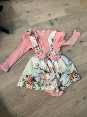 Rosa bodyset med blommig kjol - Supersött set med långärmad rosa ribbad body med volangdetaljer och matchande blommig kjol med hängslen. Kjolen har ett vackert mönster i beige, grönt och orange med stora rosor. Perfekt för dig som gillar fin och stilig stil. För 1xirka -2,5 år 