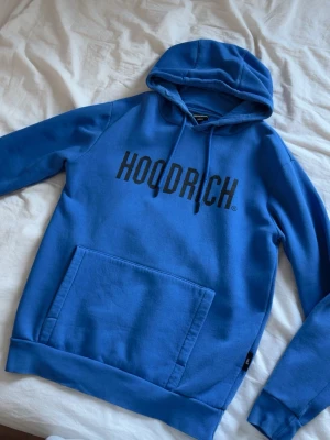 Blå hoodie från Hoodrich - Snygg blå hoodie från Hoodrich med svart logga tryckt över bröstet. Klassisk känguruficka framtill, dragsko i huvan och ribbade muddar vid ärmslut och nederkant. Tillverkad i mjukt bomullsmaterial som är skönt att ha på sig.
