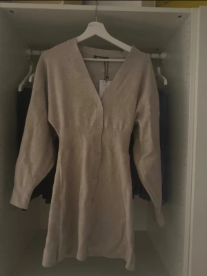 Beige lång kofta med knappar - Lång, beige kofta med v-ringning och knäppning framtill. Koftan har långa ärmar och en rak, något figurnära passform. Tillverkad i mjukt stickat material som känns skönt mot huden. Perfekt att slänga över en topp för en avslappnad look.